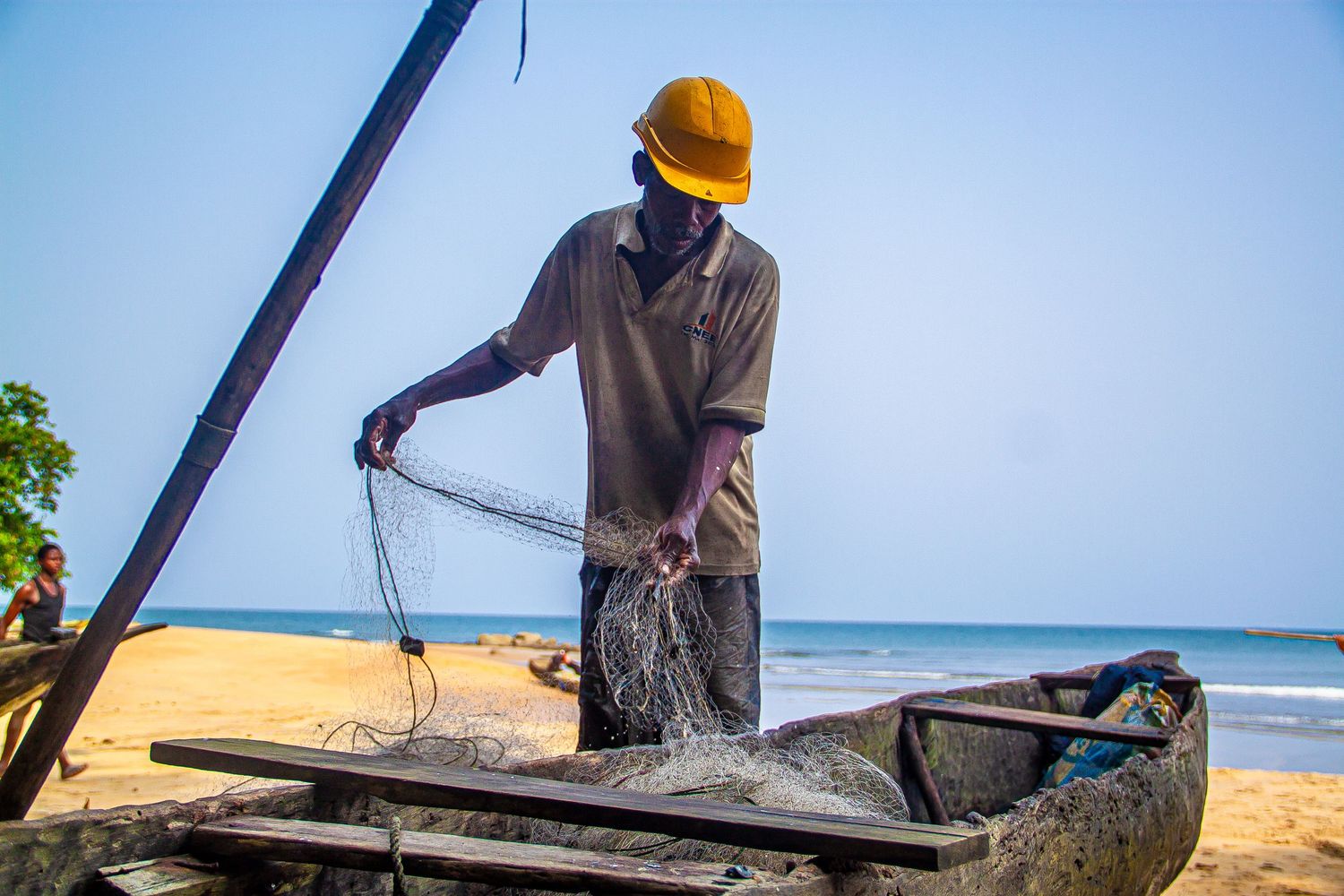 African fisherman in yellow hard hat sorting fishing nets on a wooden boat on a sunny sandy beach.