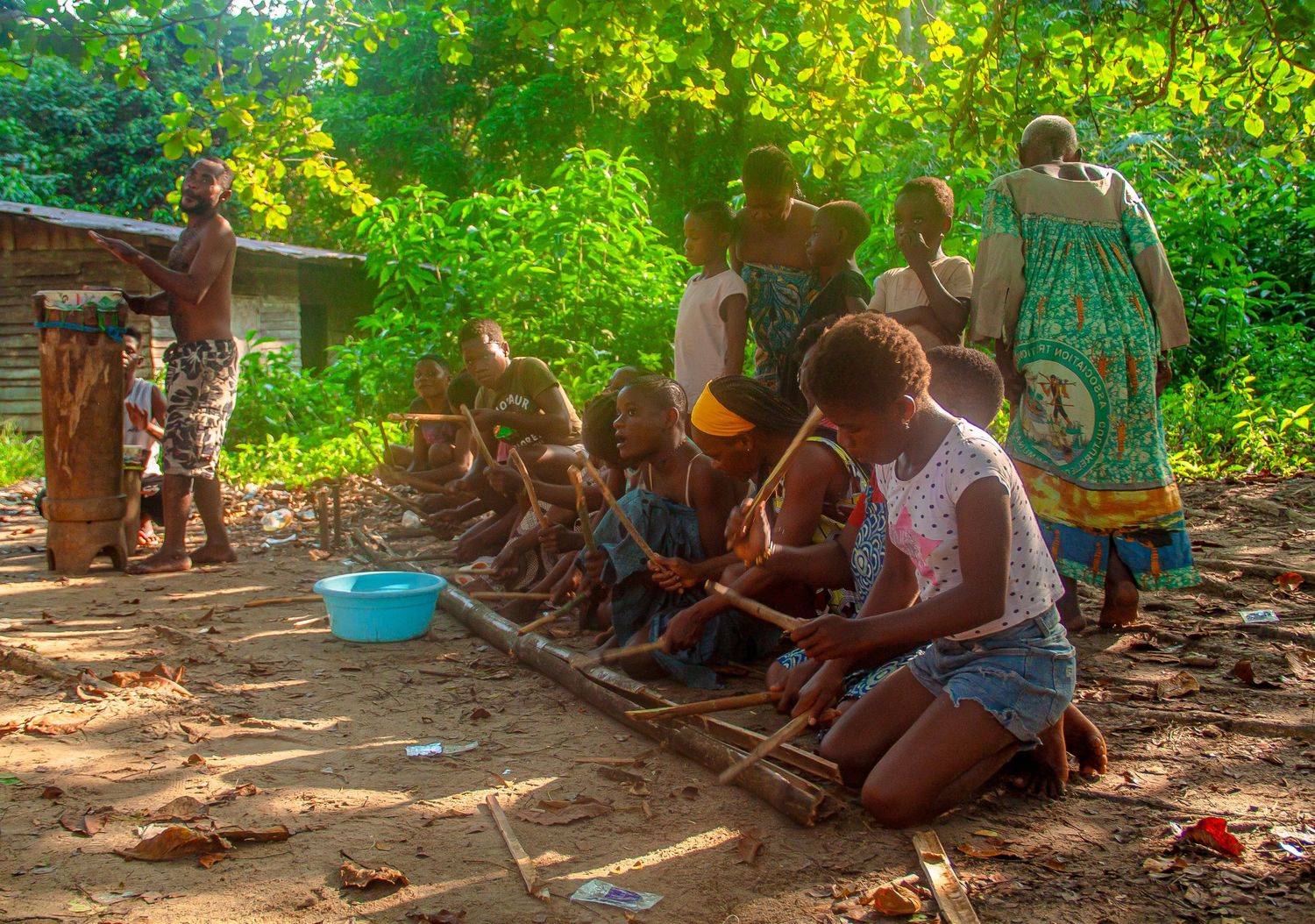 African women and children kneeling outdoors in a village clearing surrounded by lush green trees.