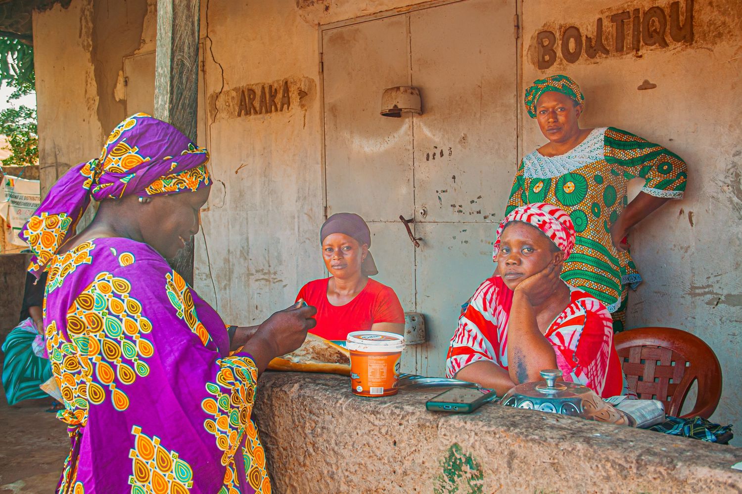 Four women in vibrant traditional clothing socializing outside a small boutique shop in West Africa.