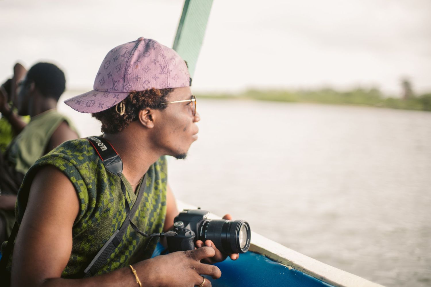 A young man wearing a pink cap holds a camera while sitting on a boat overlooking a river.