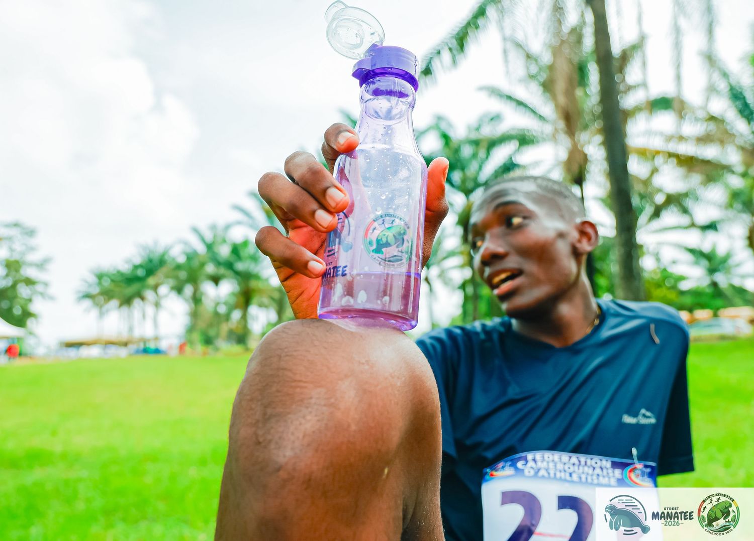 Athlete pouring water from purple bottle on neck during outdoor race event.