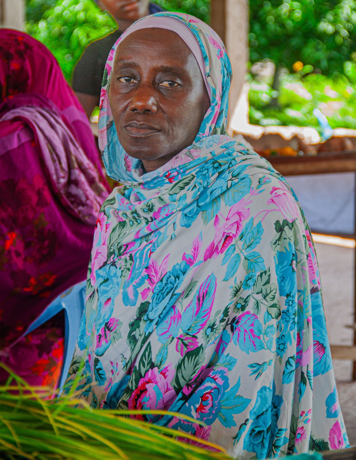 Senegalese woman wearing a floral blue hijab sitting outdoors in a lush green setting.