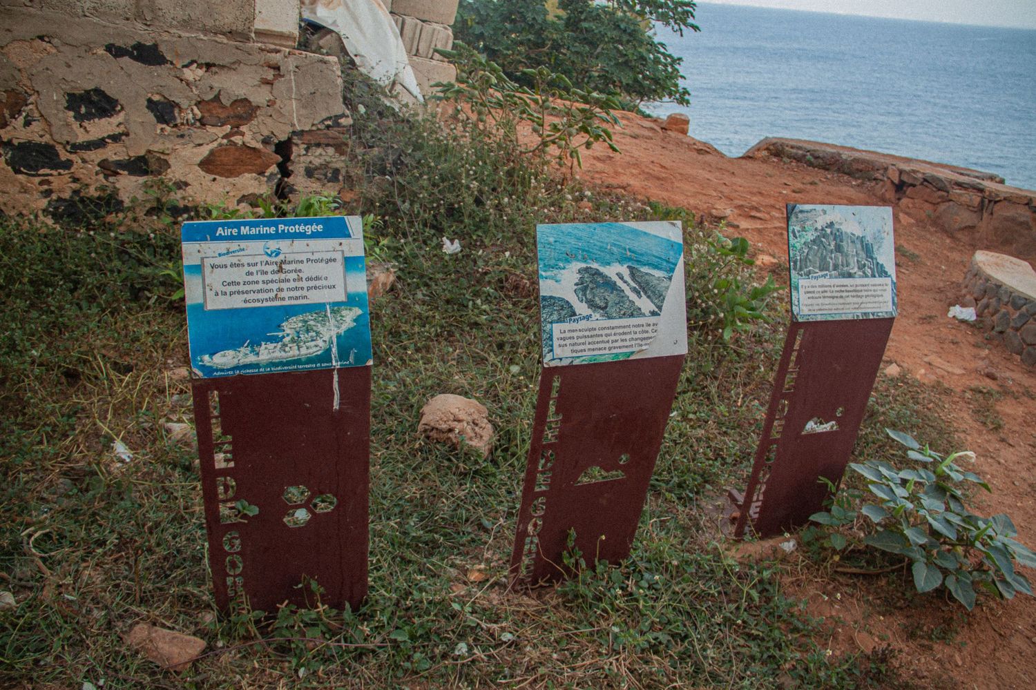Three rusty informational sign posts with tourist maps near a red coastal cliff overlooking the ocean.