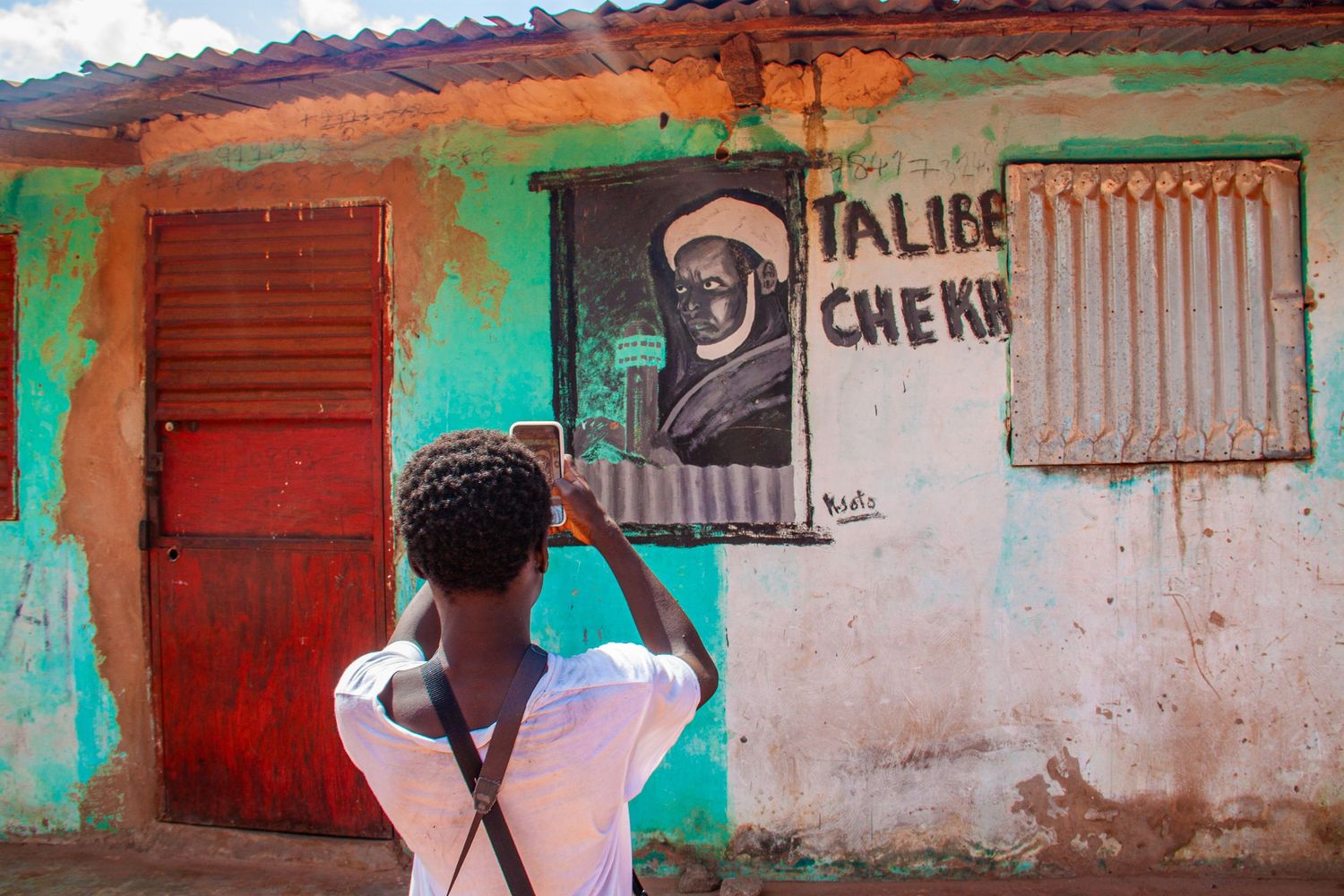 A man photographs a painted mural of Taliibe Cheikh on a vibrant turquoise wall in Senegal.