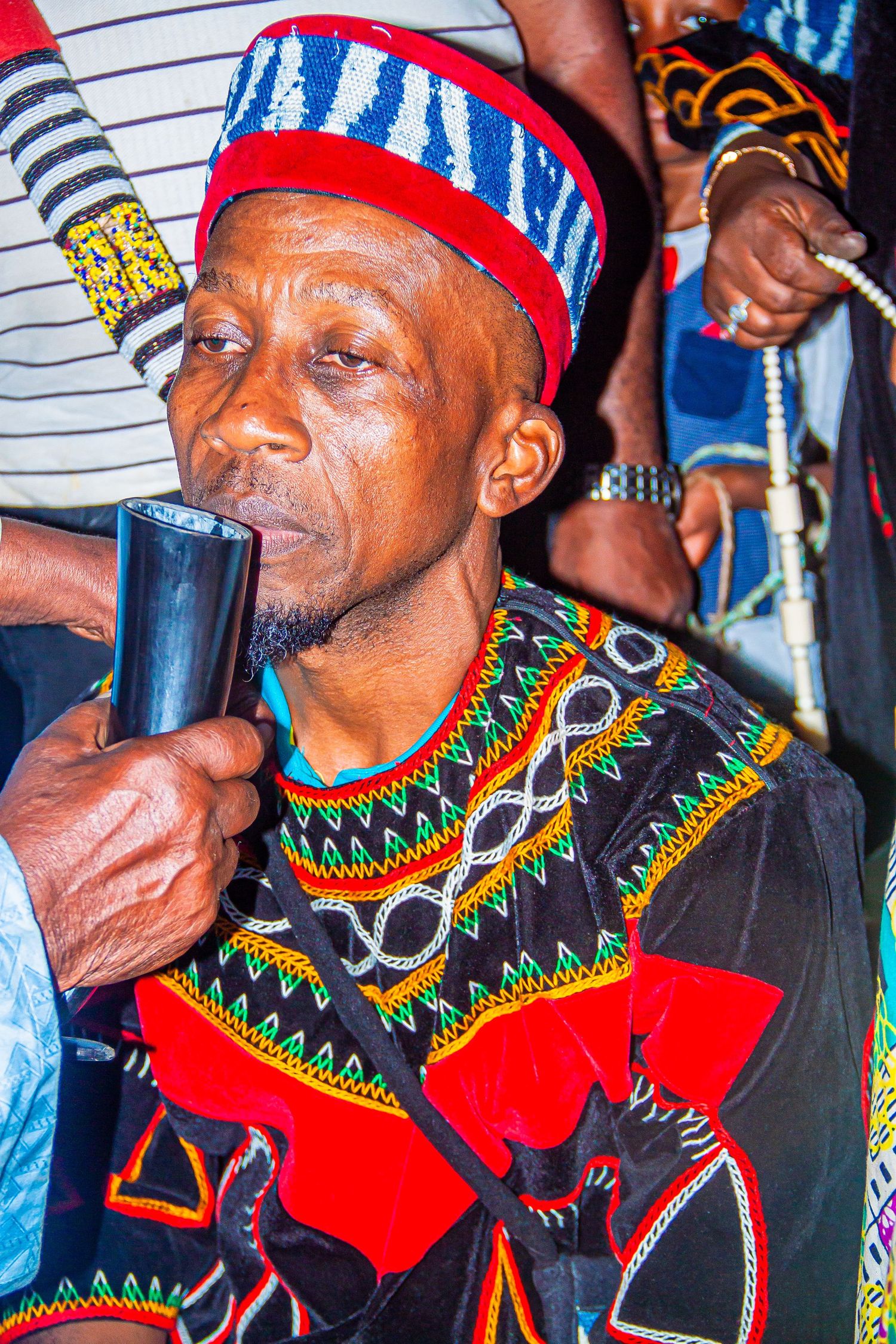 African man in traditional colorful embroidered attire and beaded hat playing a traditional horn instrument.