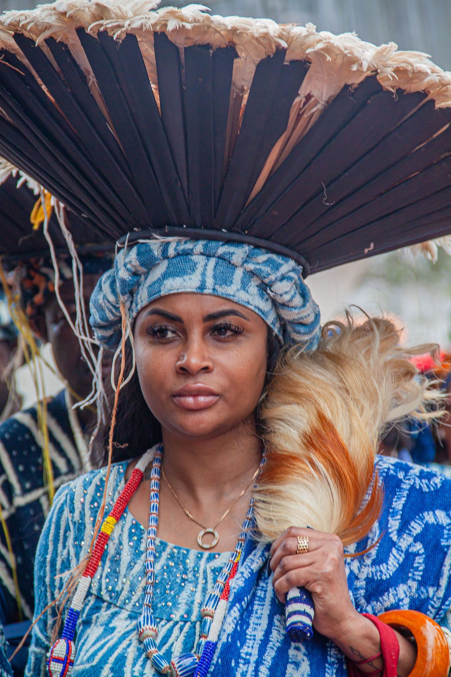 African woman in blue traditional attire with large dark feathered hat and beaded jewelry at cultural festival.