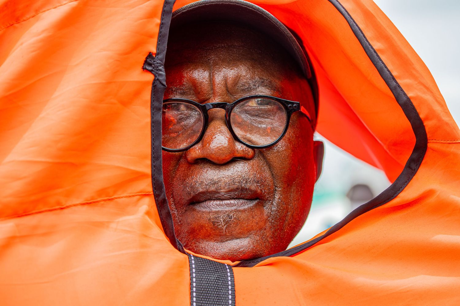 Elderly man peering through an orange hooded jacket with a serious expression on his face.