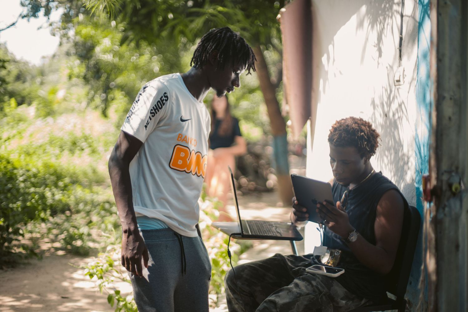 Two young men collaborate outdoors, one standing while the other works on a laptop in the shade.