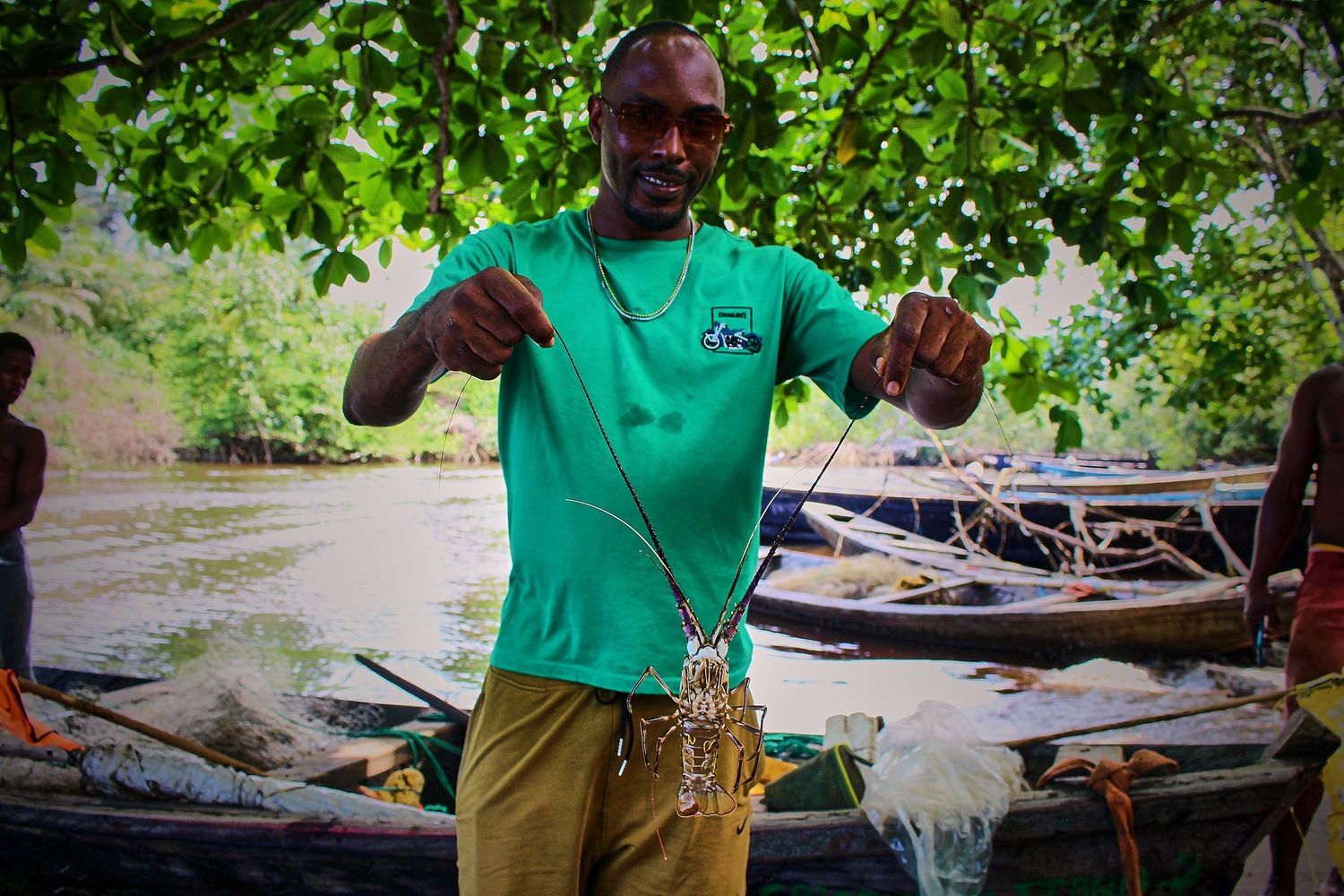 A smiling man in a green shirt holds up a lobster catch beside wooden canoes near a river.