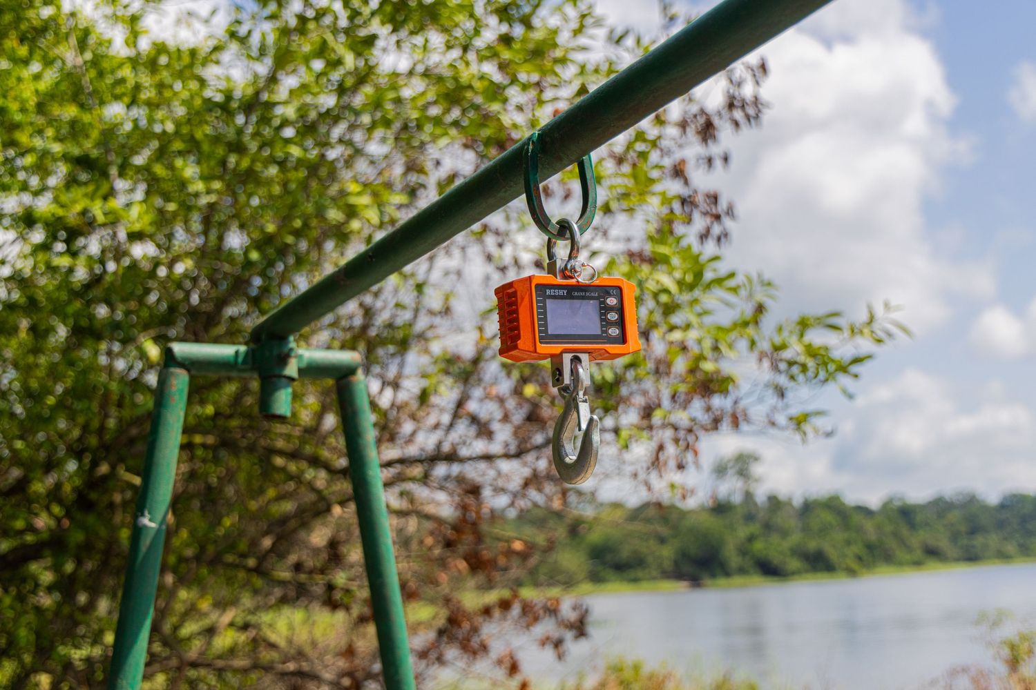 Orange digital hanging scale suspended from a metal frame outdoors near a tropical river environment.