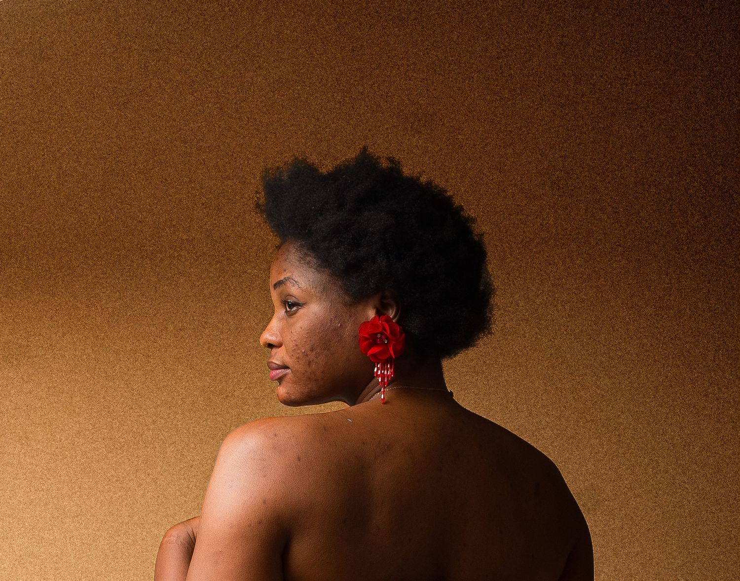 Black woman with natural afro hair and red floral earrings posing with bare back against brown background.