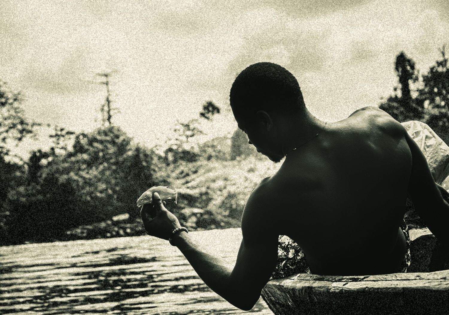 Shirtless man sitting by river holding small object in vintage black and white photo.