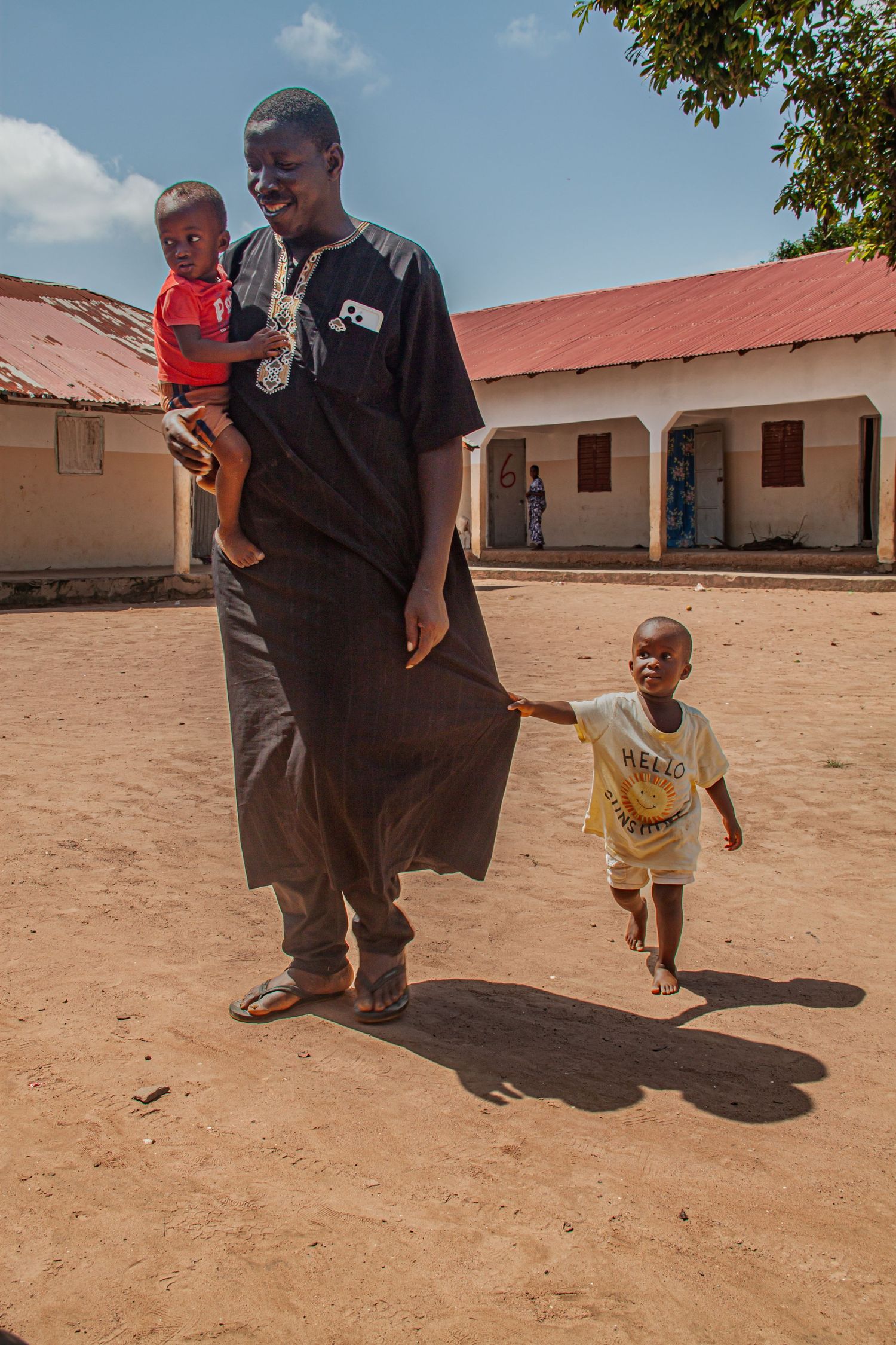 African man in traditional black attire carrying a toddler while another child holds his clothing.
