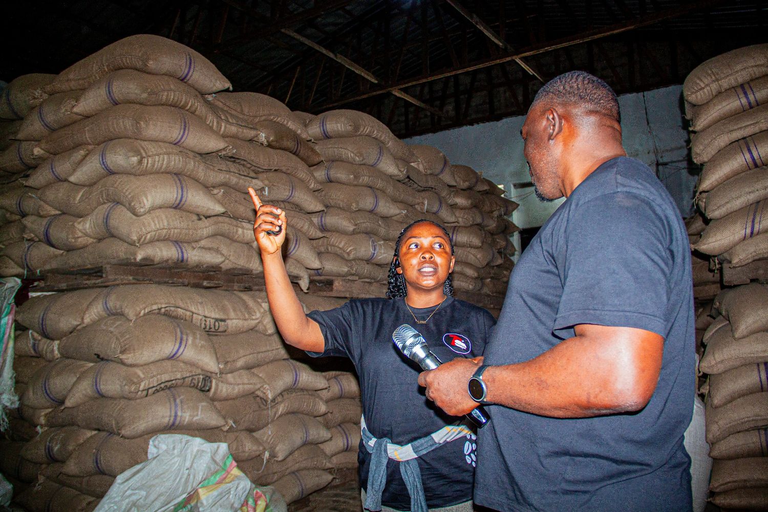 Two men in black shirts inspect large burlap sacks stacked in a warehouse storage facility.