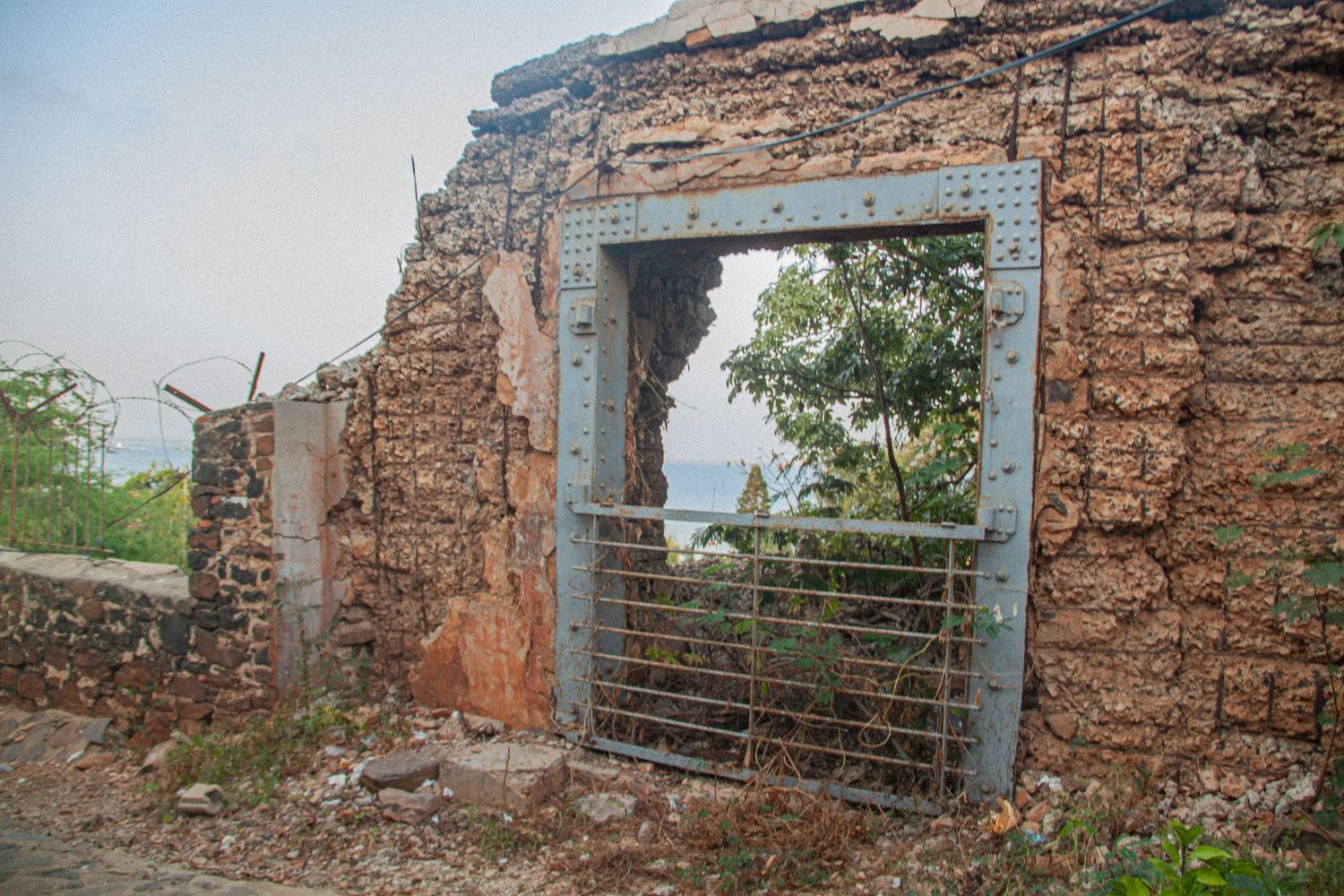 Ruins of an ancient stone building with a metal gate doorway revealing trees and ocean beyond.