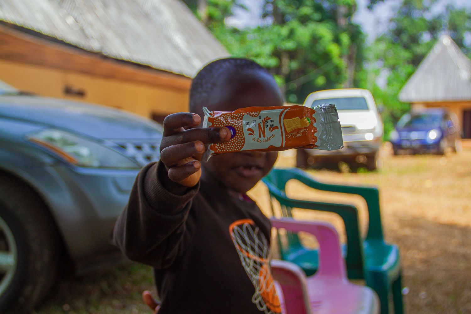 Young African child holding up a small toy or package outdoors near parked cars.