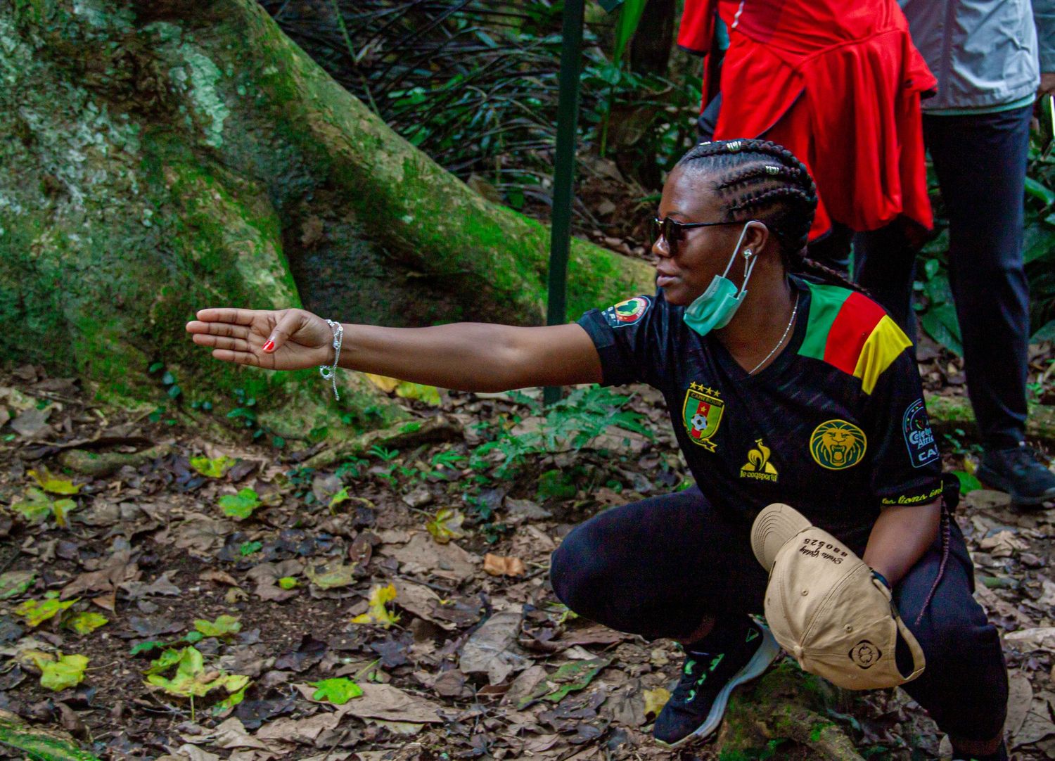 Person crouching in tropical forest, wearing Cameroon jersey and face mask, pointing at the ground.