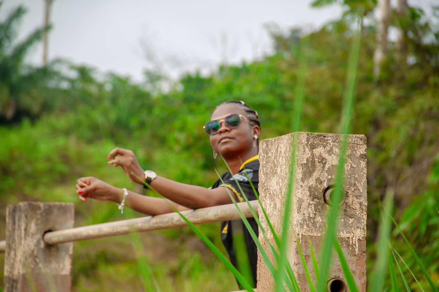 Person in sunglasses leaning on wooden gate surrounded by lush green vegetation.