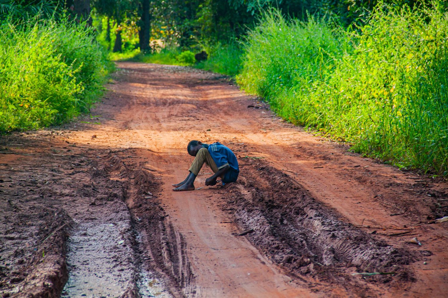 Person sitting alone curled up on muddy red dirt road surrounded by lush green vegetation.