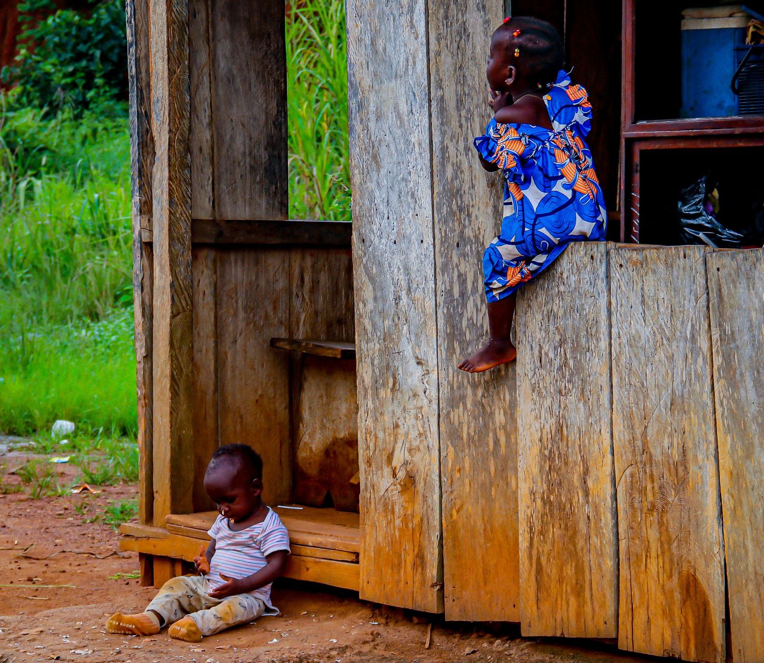 Two young African children play outside a rustic wooden building in a rural village.
