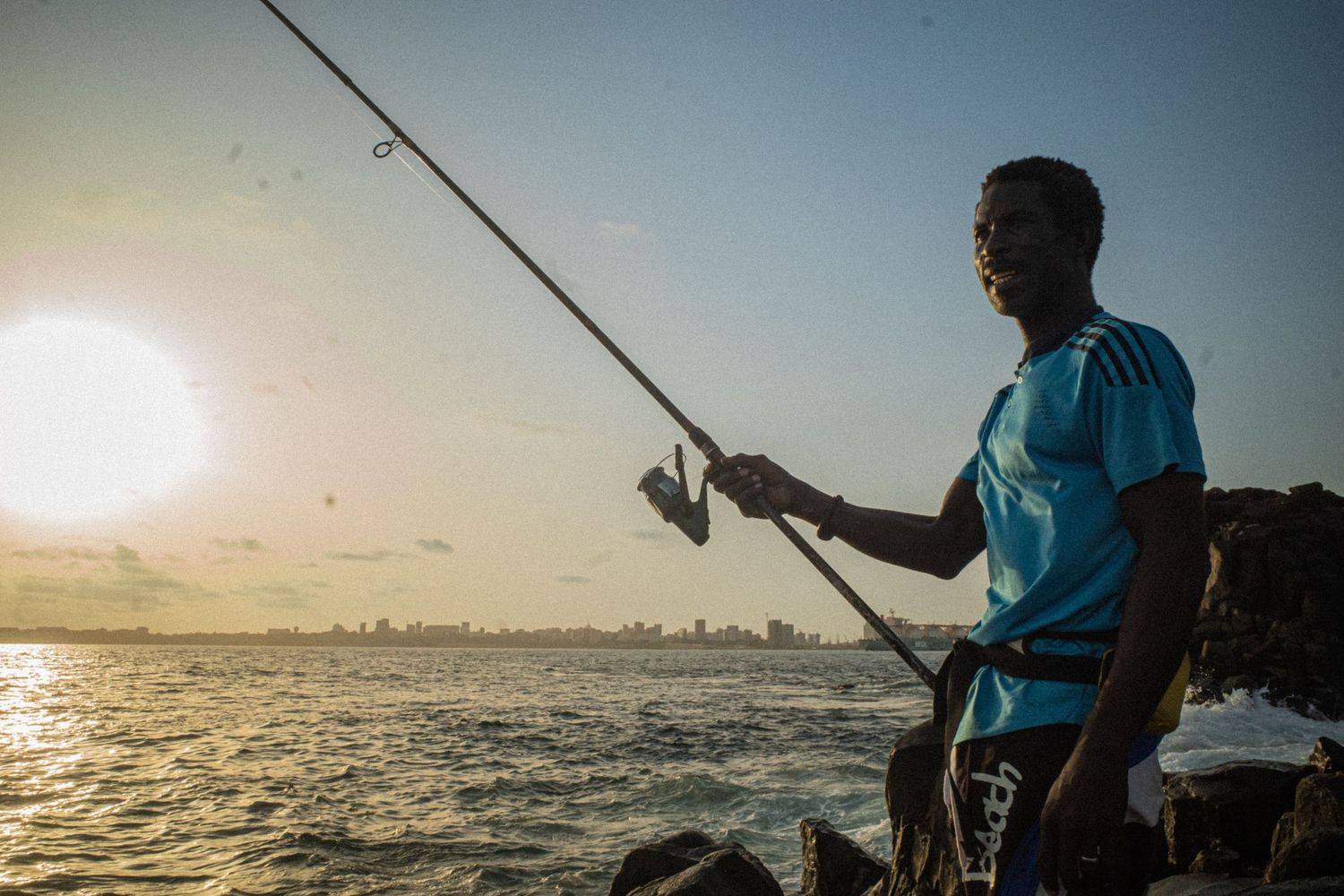 Man fishing with rod at sunset on rocky coast with city skyline in background.