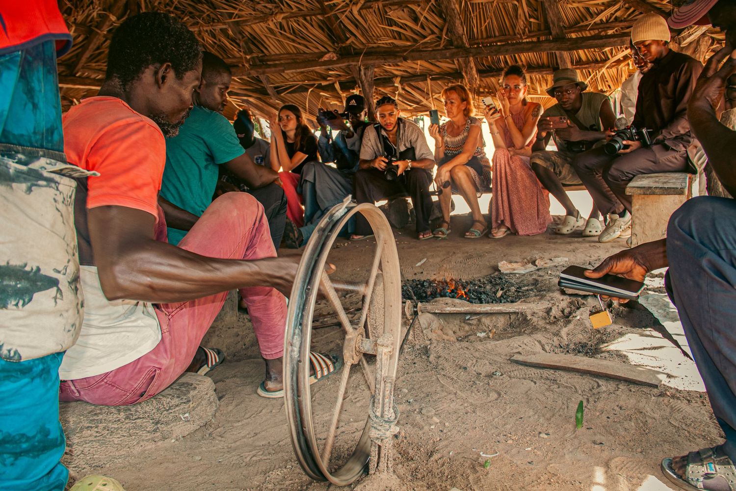 Group of African men and tourists gathered under thatched roof hut with bicycle wheel visible.