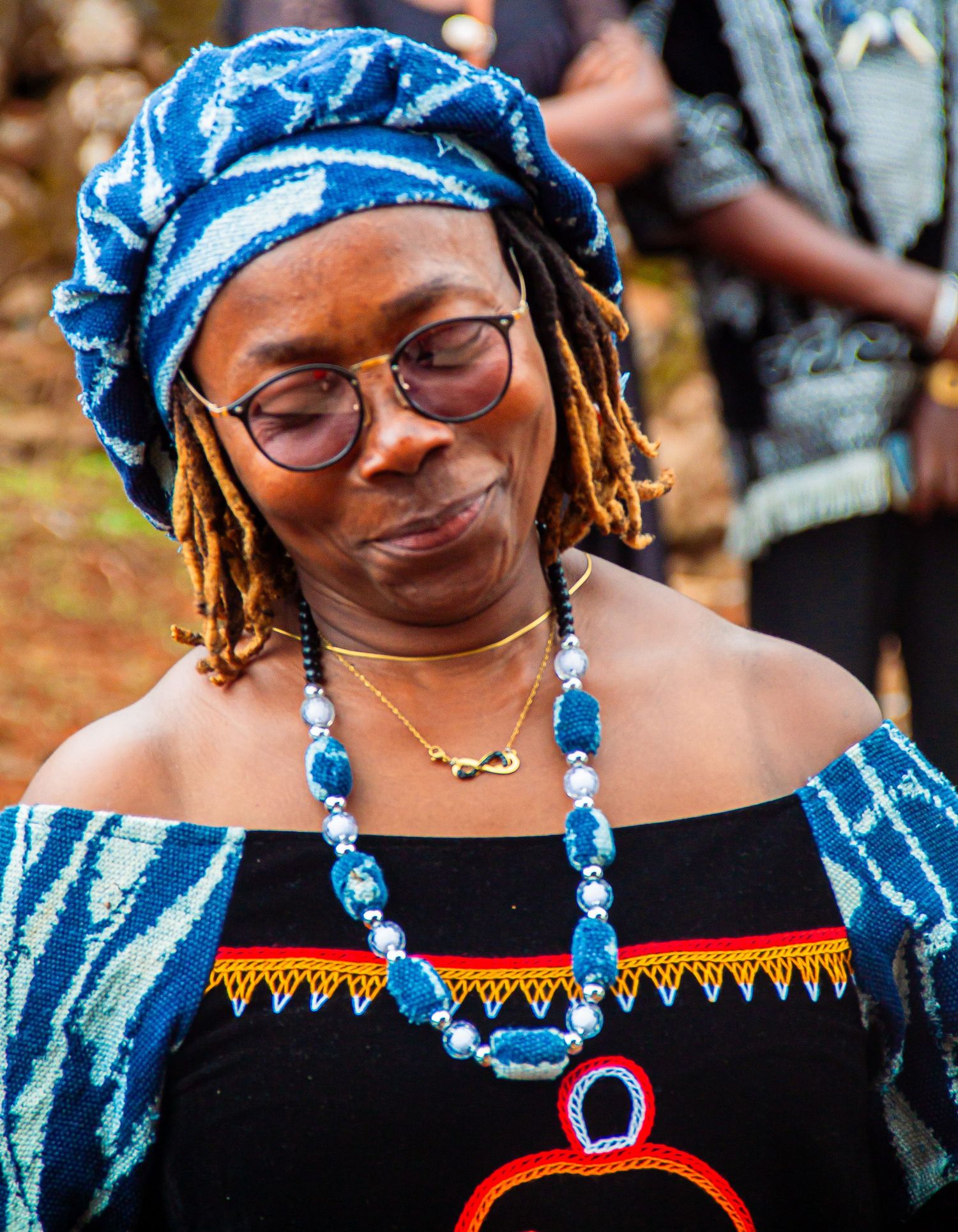 An African woman in traditional blue head wrap and colorful necklace wearing round glasses outdoors.