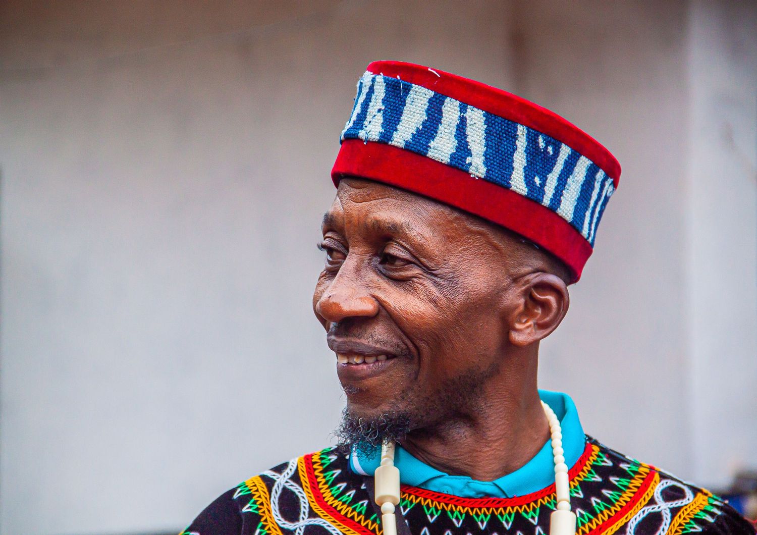 Smiling elderly African man in colorful traditional attire wearing a red and blue patterned cap.