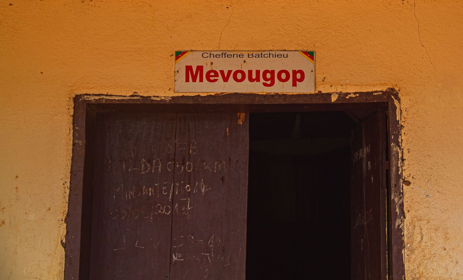 Sign reading Mevougop on yellow wall above wooden door in African village building.