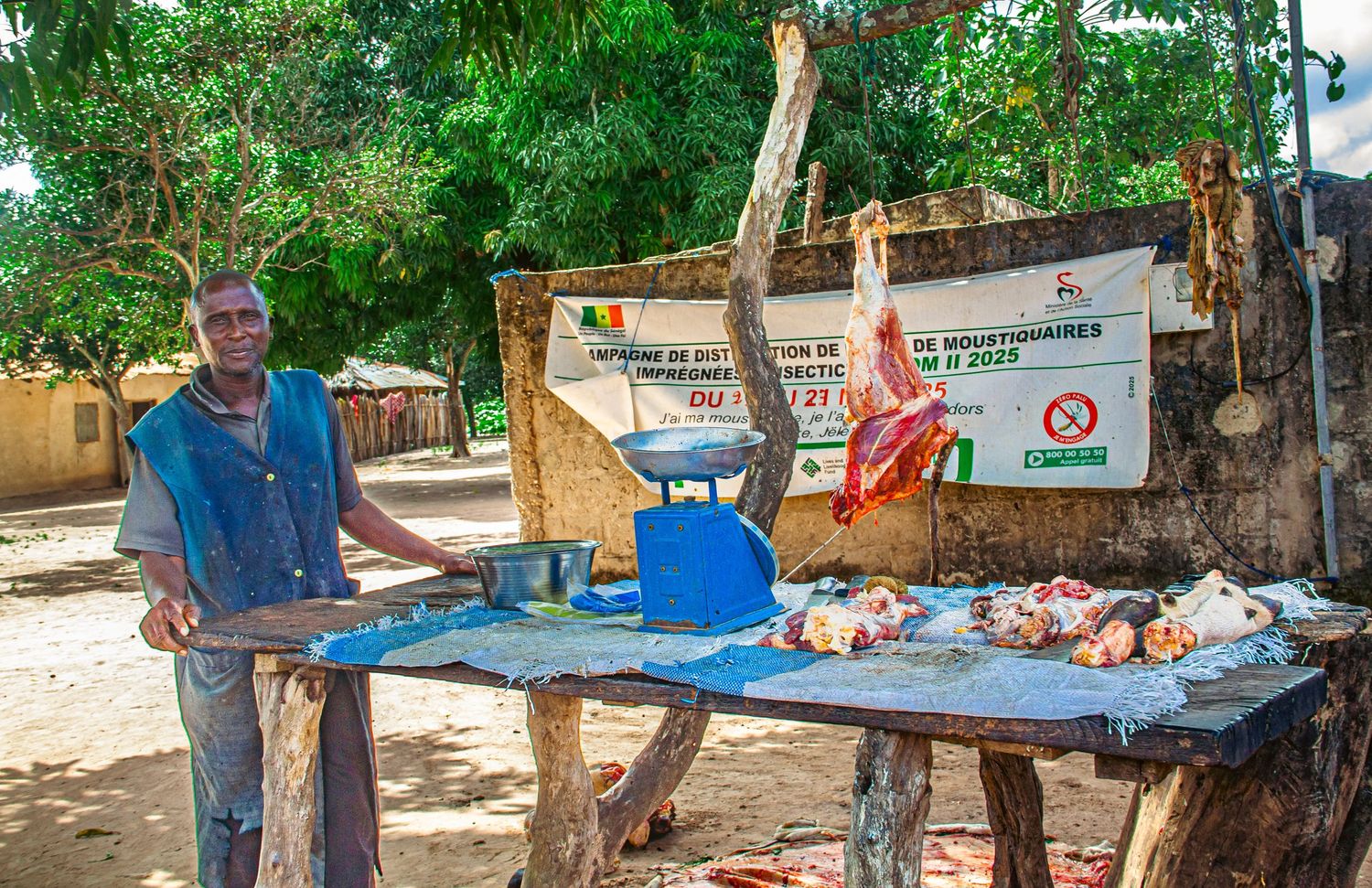 A man sells fresh meat at an outdoor market stall in a rural West African village setting.