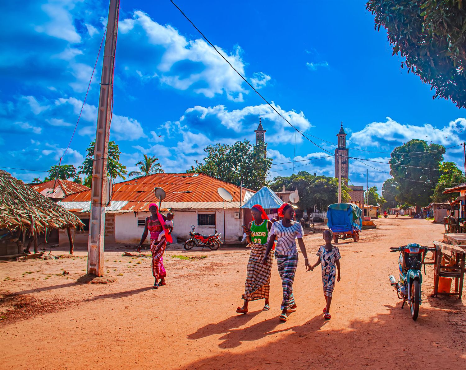 Colorful African village street scene with women and children walking on red dirt road under blue sky.
