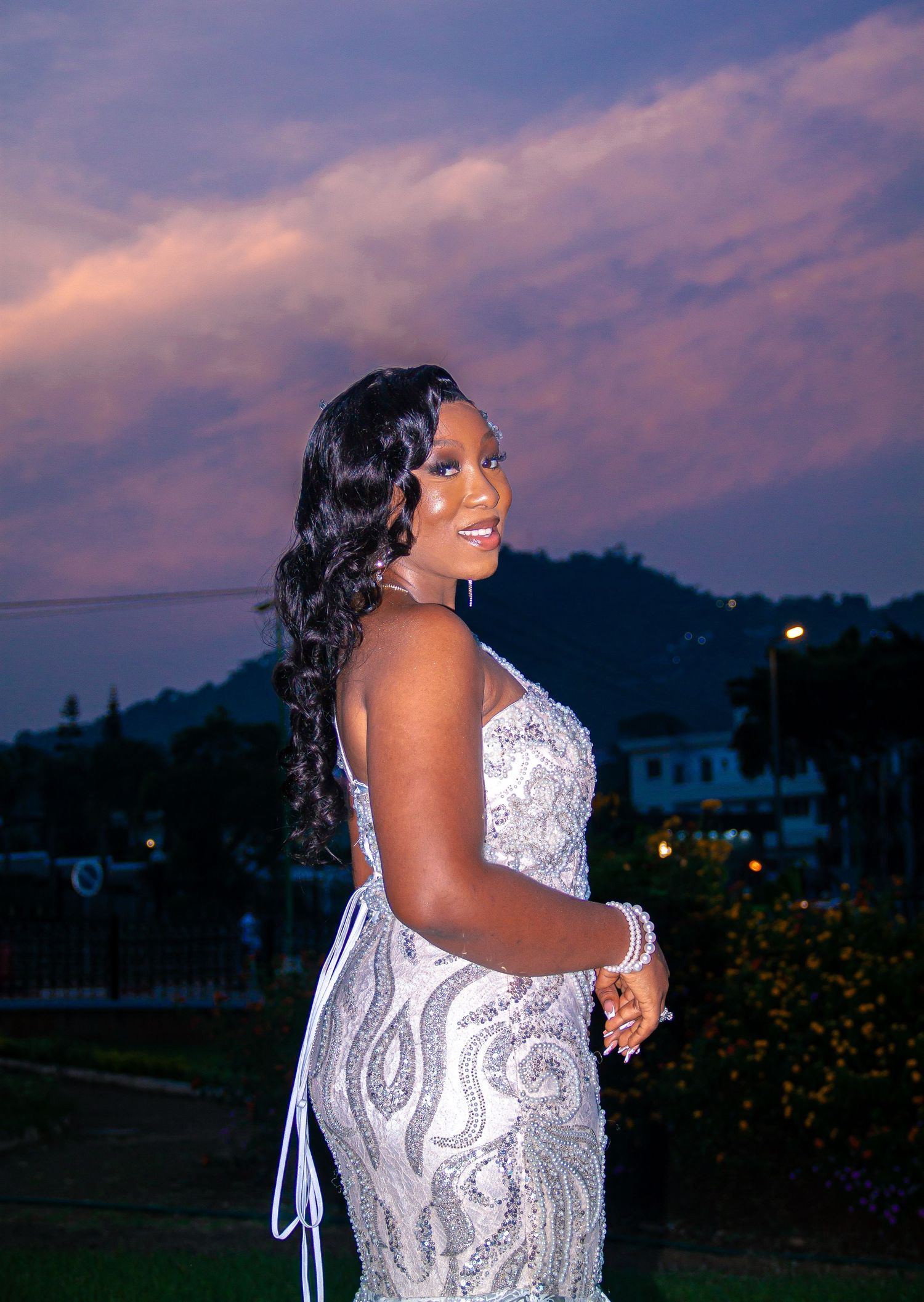 Woman in elegant beaded white gown posing at sunset with purple sky backdrop.