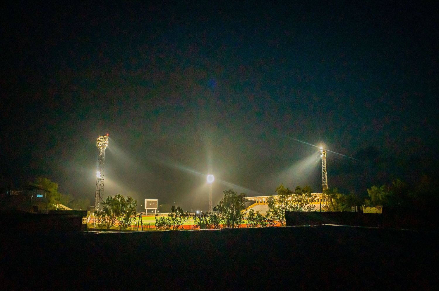 A sports field illuminated by bright floodlights at night surrounded by tropical vegetation.