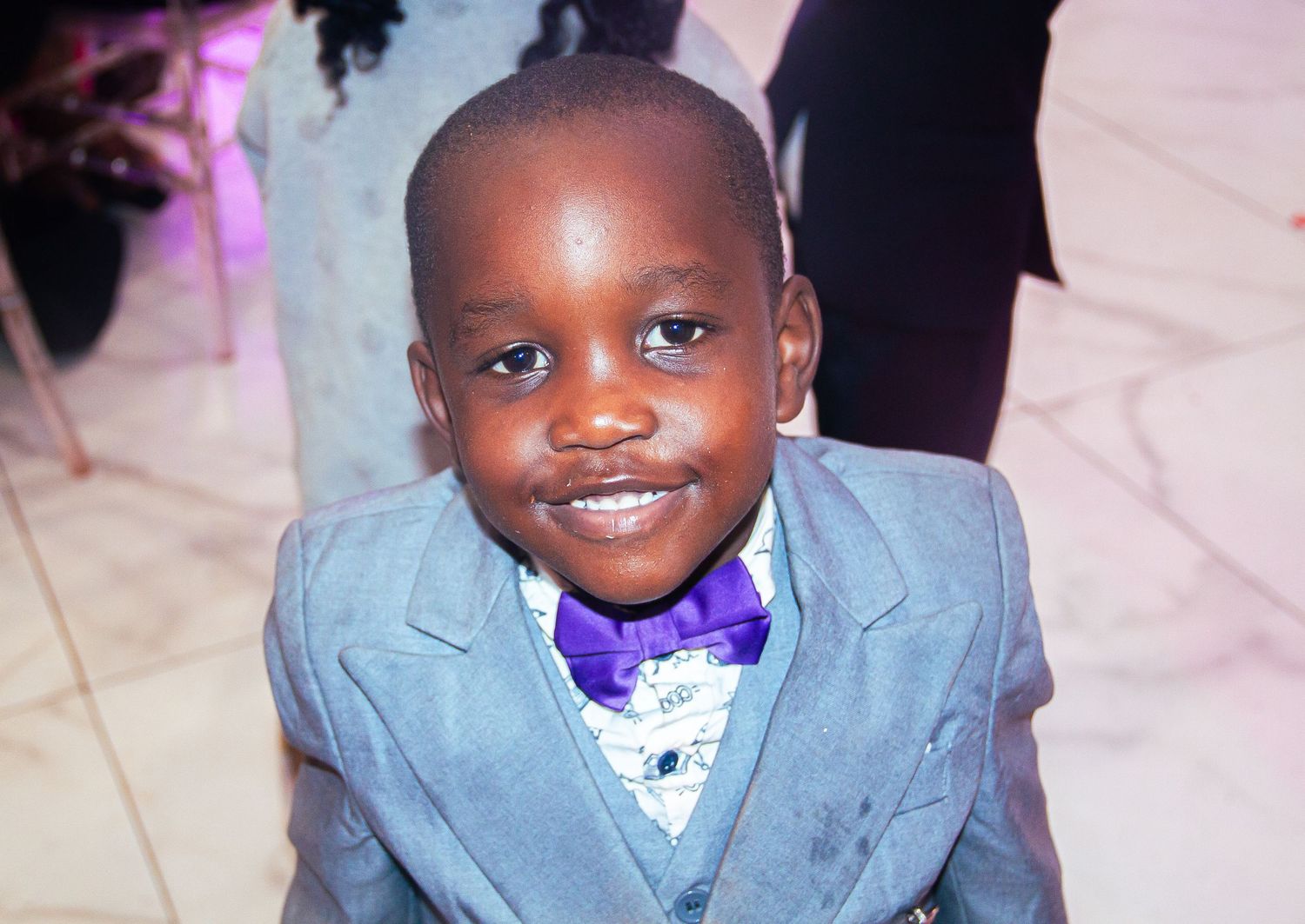 A young African boy in a gray suit and purple bow tie smiles at a formal event.