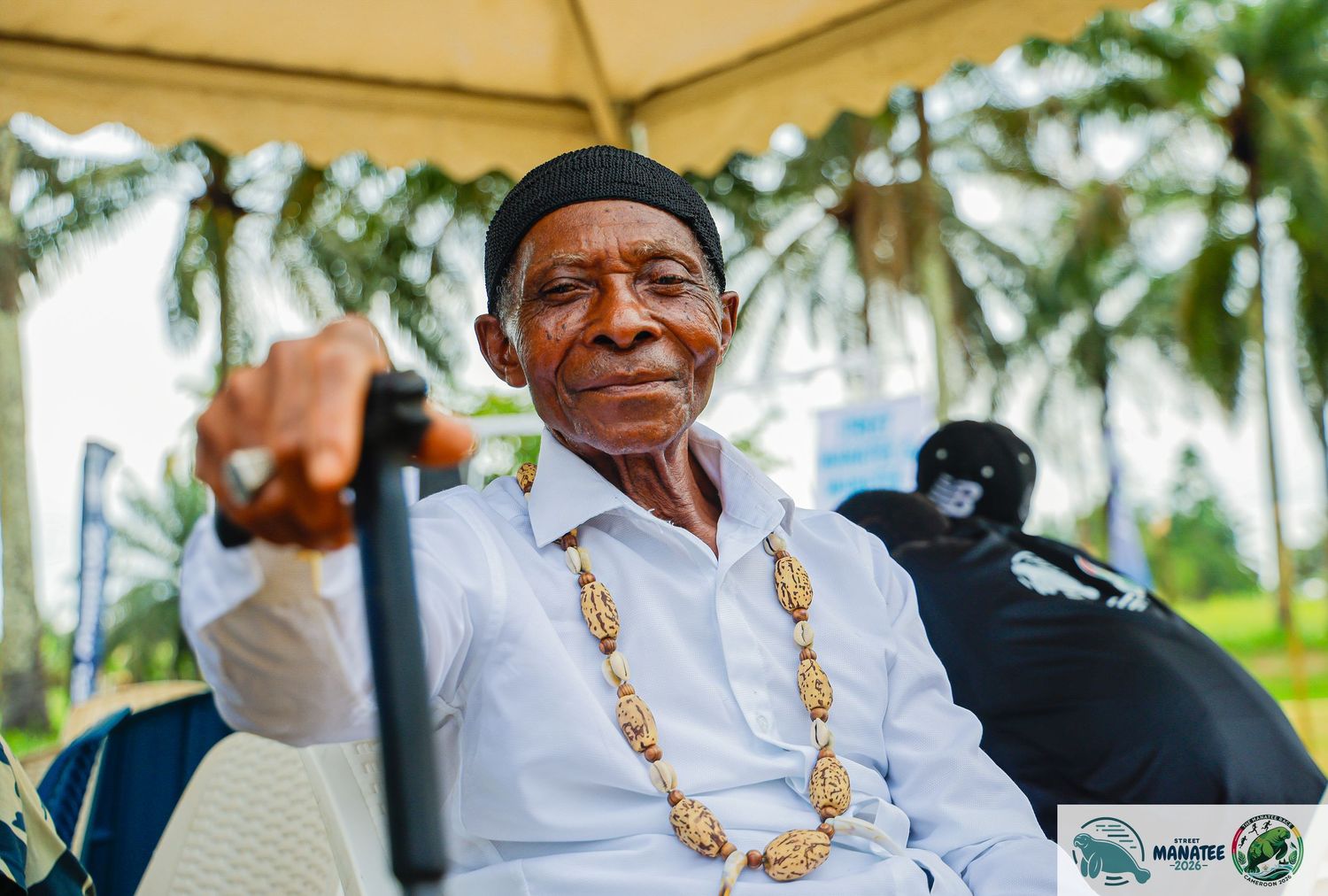 Elderly man in white shirt with beaded necklace seated outdoors smiling.
