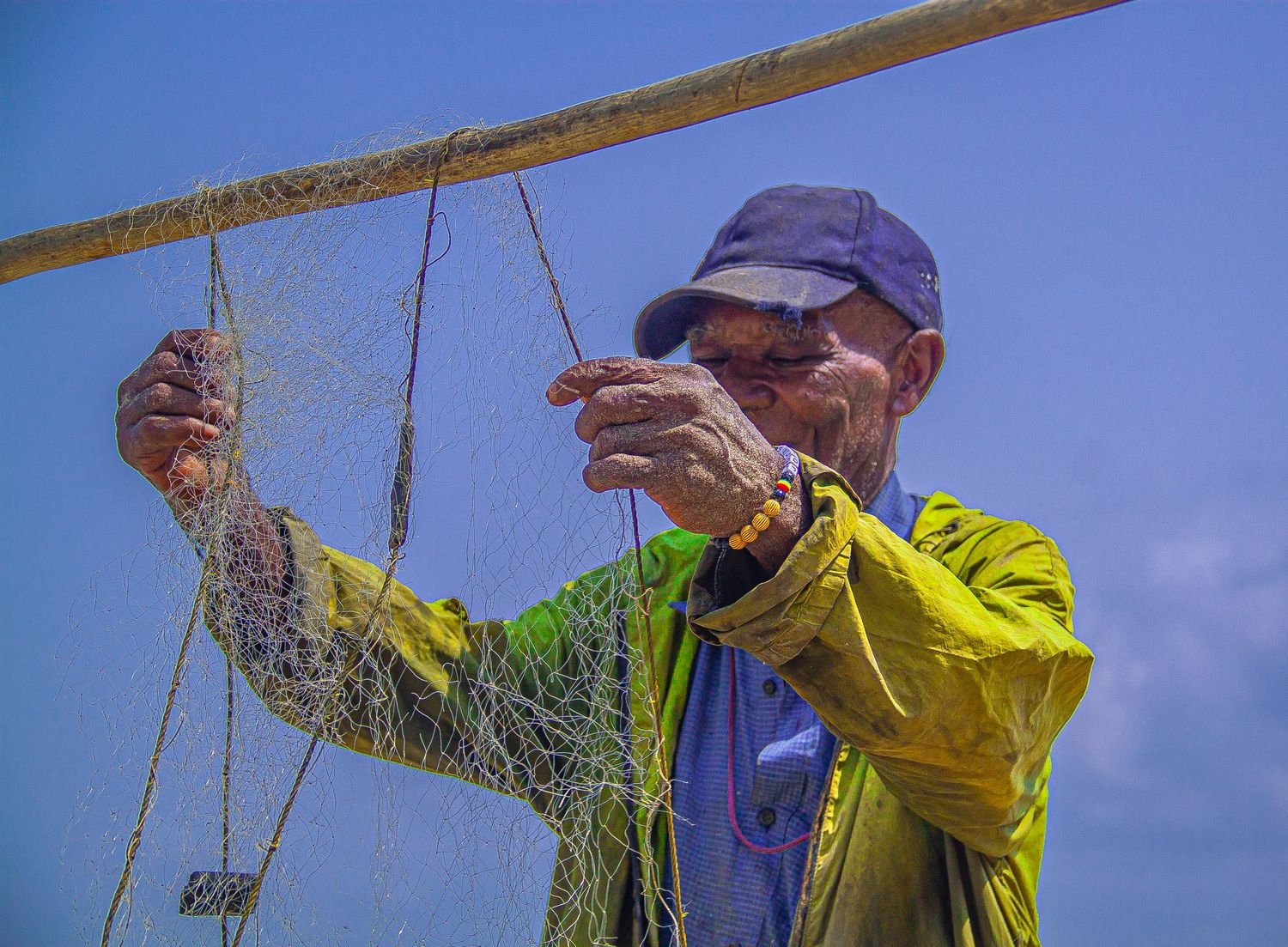 Elderly man in yellow jacket and blue cap repairs fishing net against a clear blue sky.
