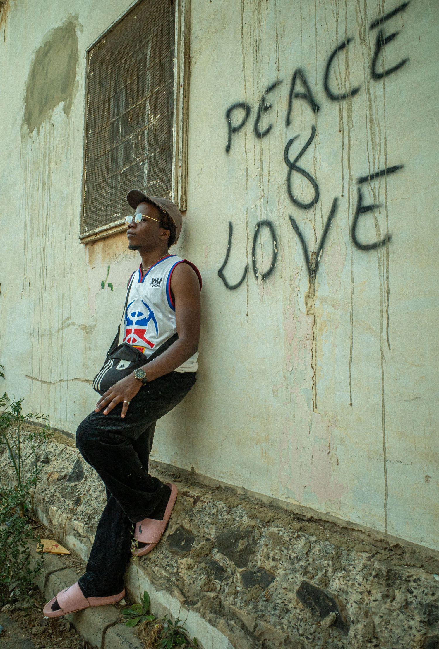 A young man leans against a weathered wall with Peace and Love graffiti written in bold black letters.