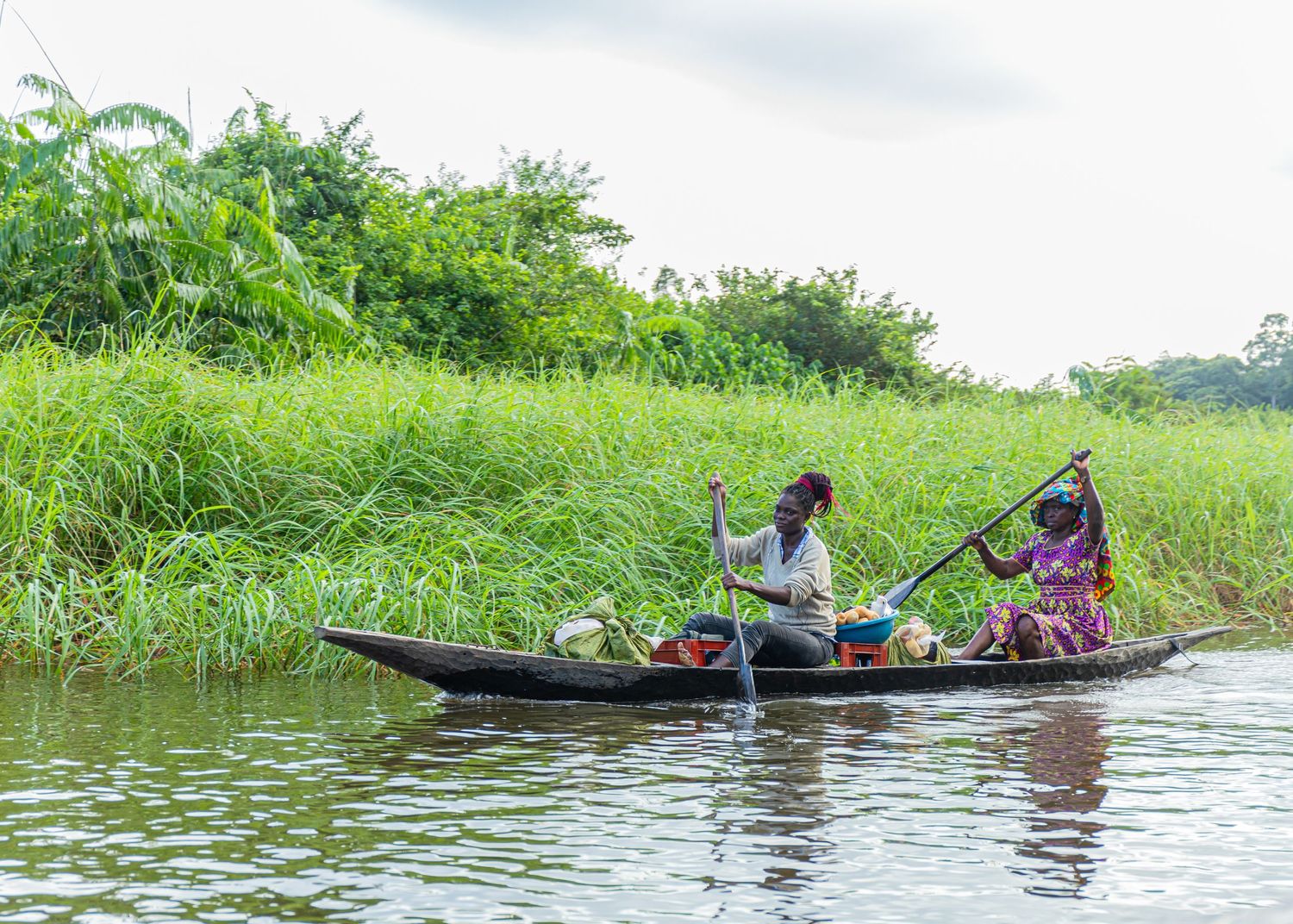 Two people rowing a wooden canoe loaded with goods along a green tropical African waterway.