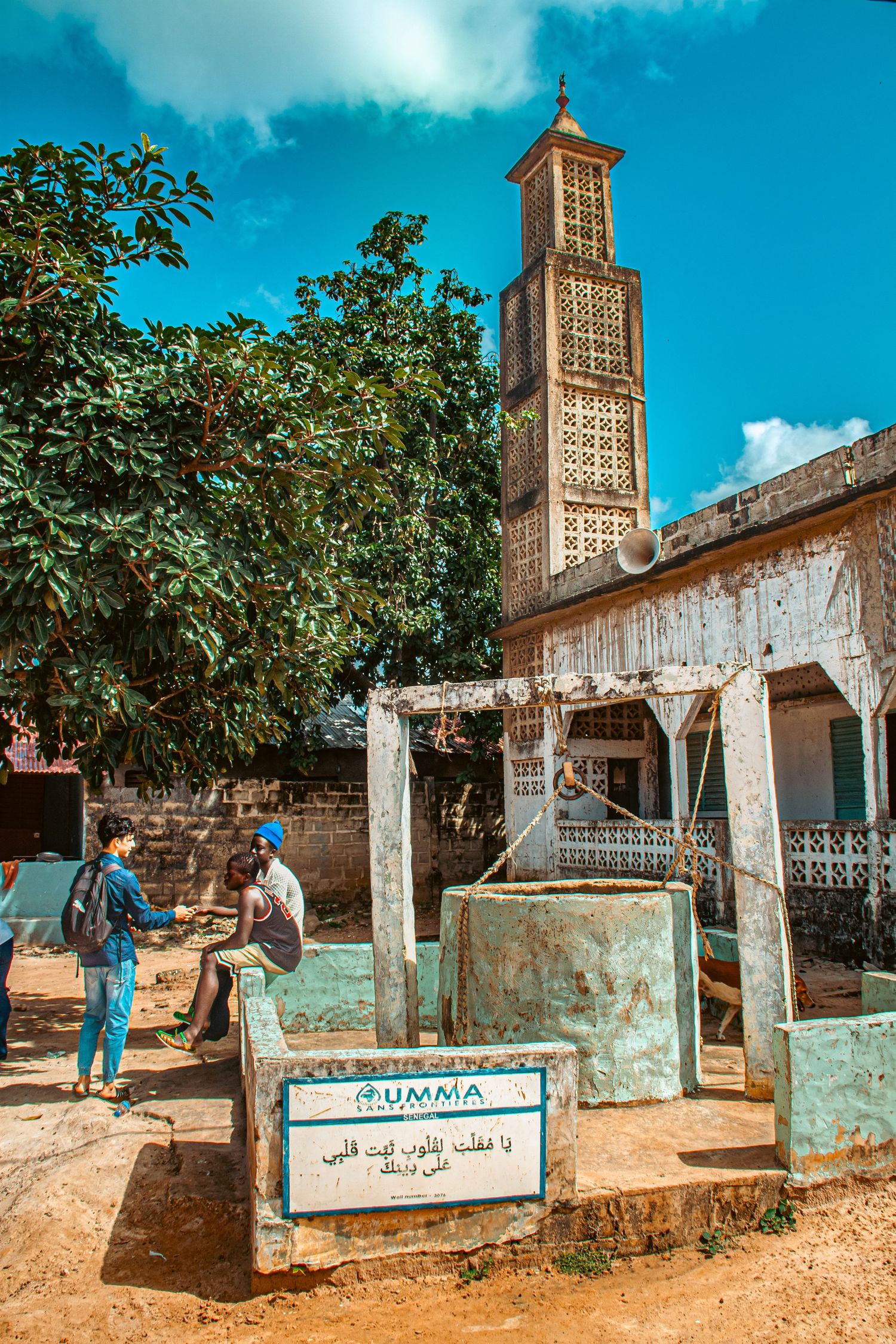 A mosque minaret rises above trees near a traditional well with Arabic inscription in a West African village.