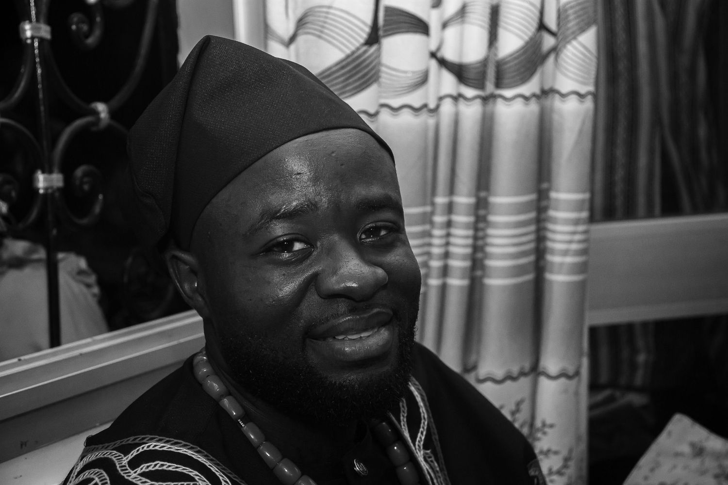 Smiling African man in traditional black embroidered cap and attire in black and white photo.