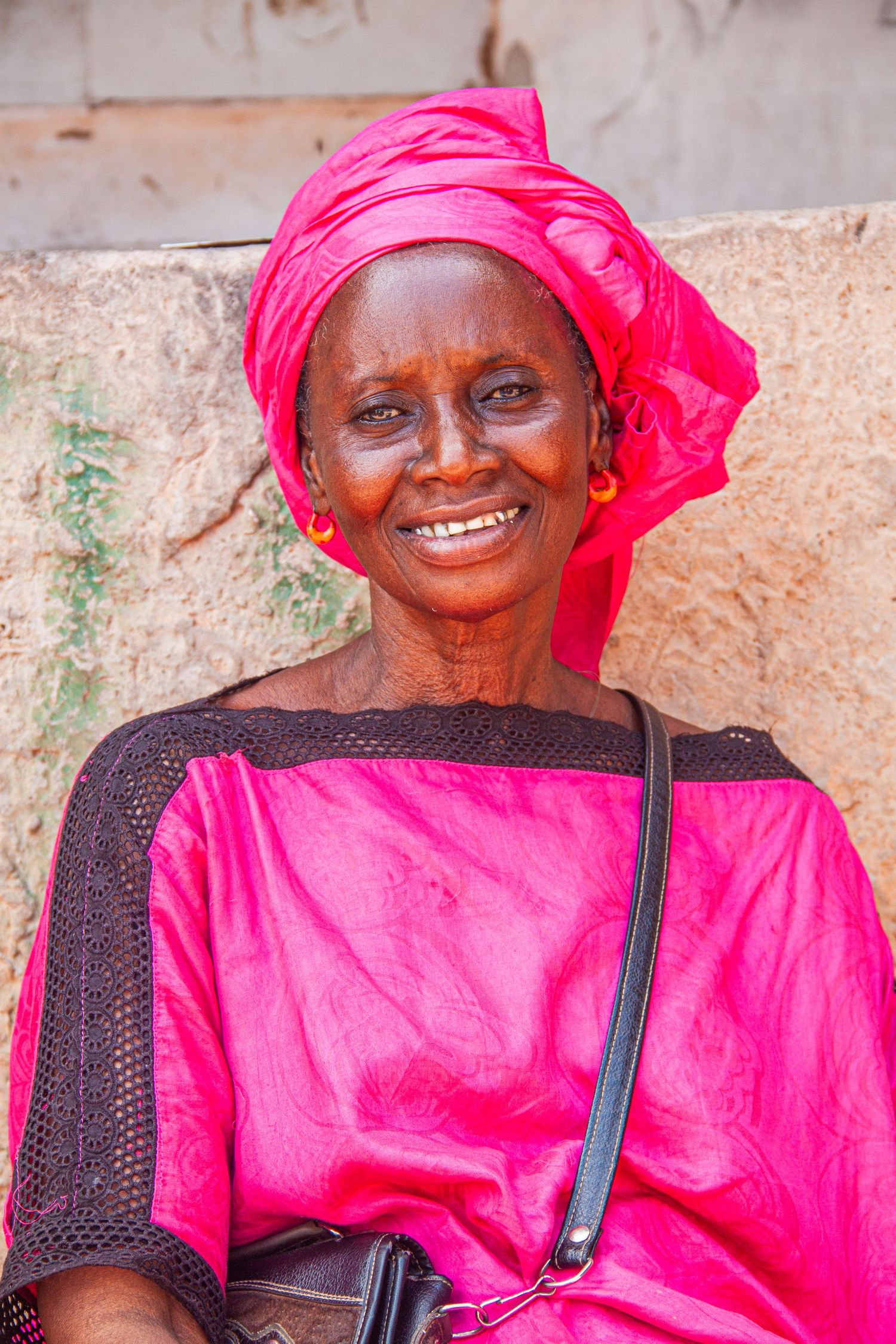 Smiling elderly Senegalese woman wearing vibrant pink traditional dress and matching headwrap.