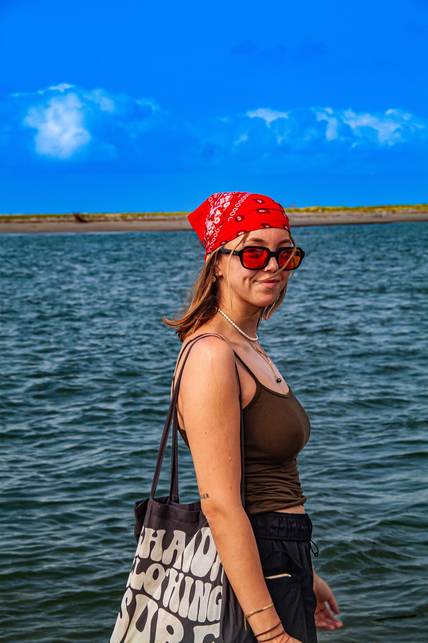 Young woman in red bandana and sunglasses posing by a lake with blue sky background.
