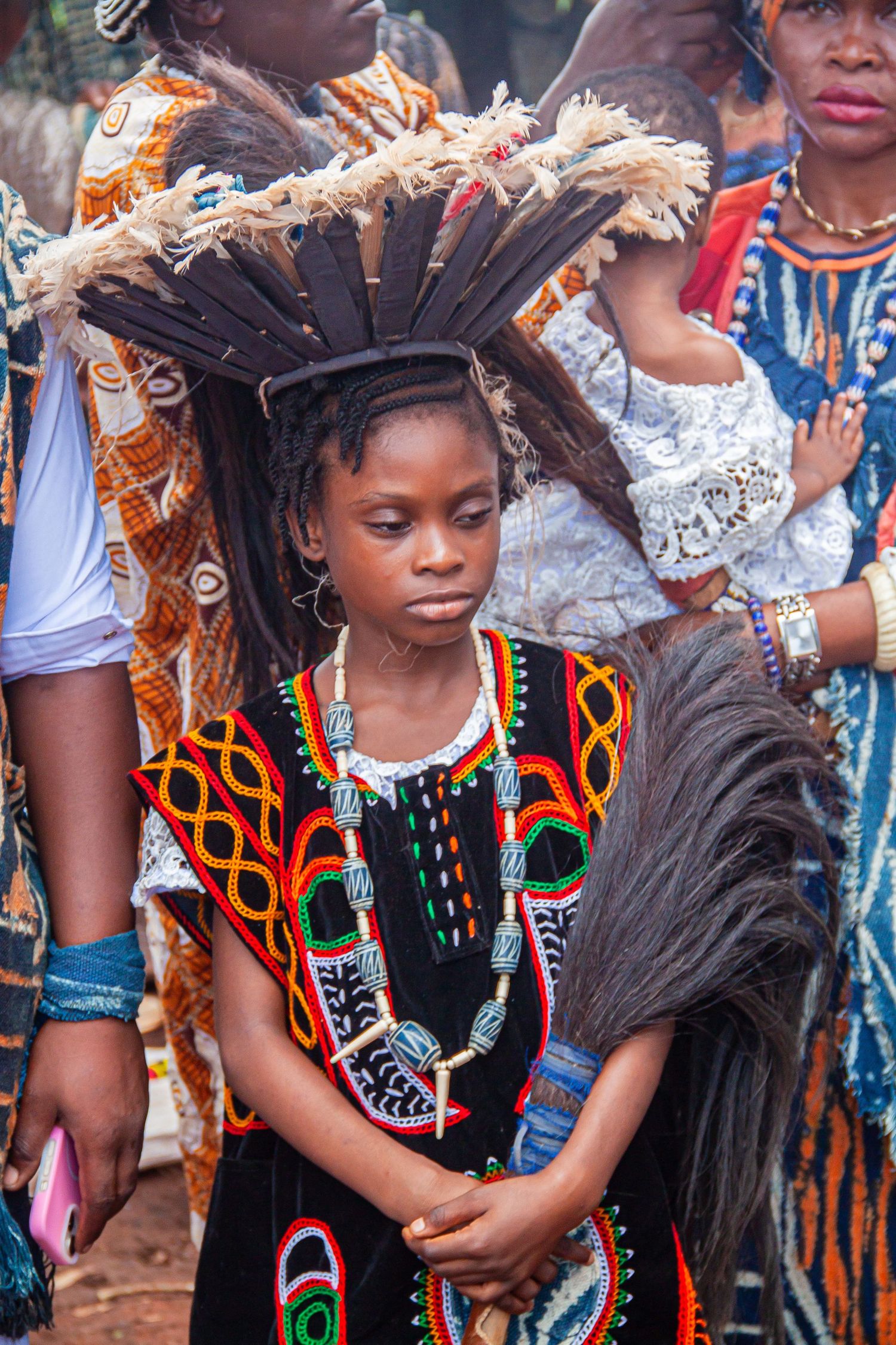 Young African girl in vibrant traditional beaded costume wearing a large feathered headdress at festival.
