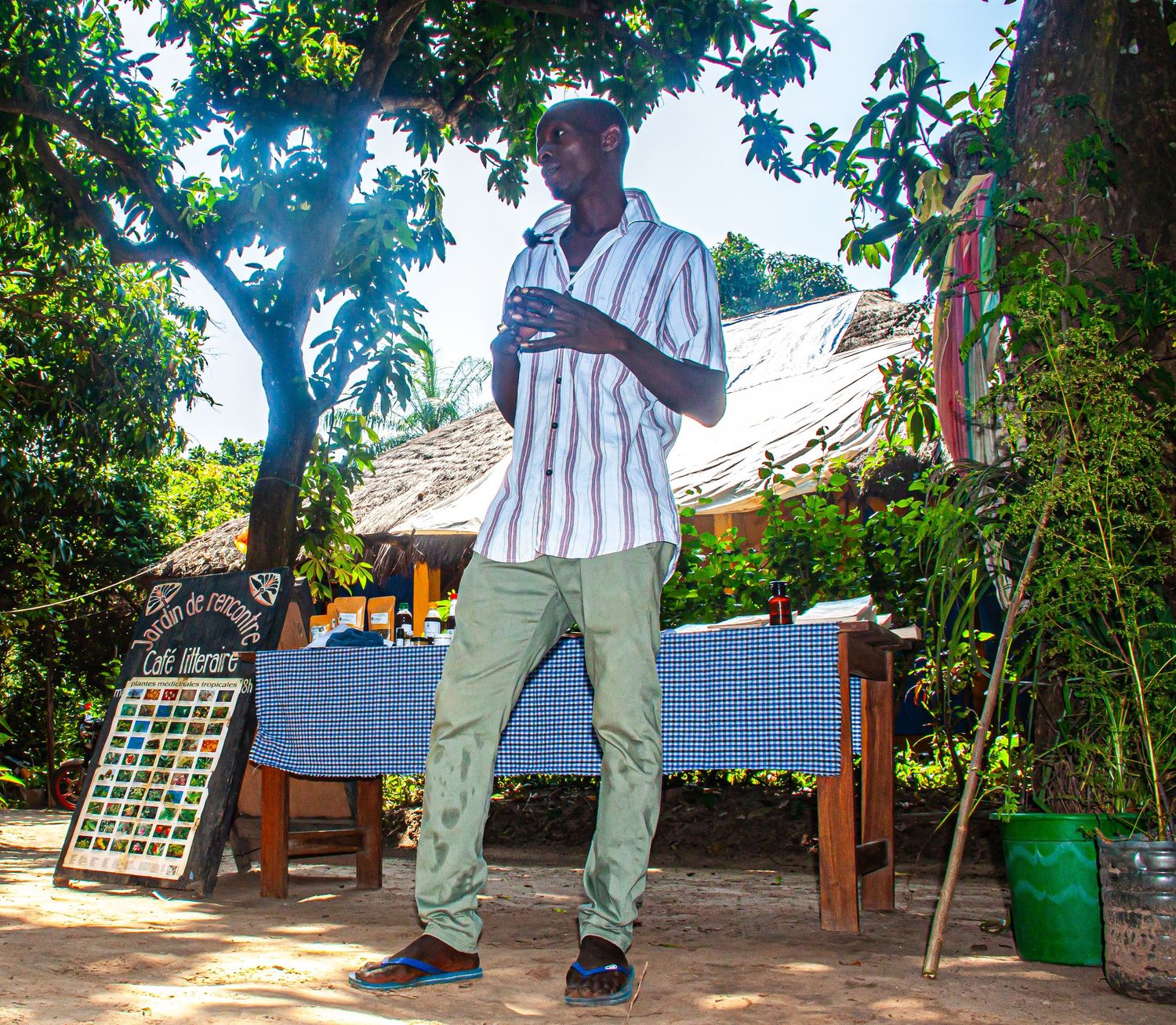 Tall African man in striped shirt speaking outdoors under shady trees in a village.