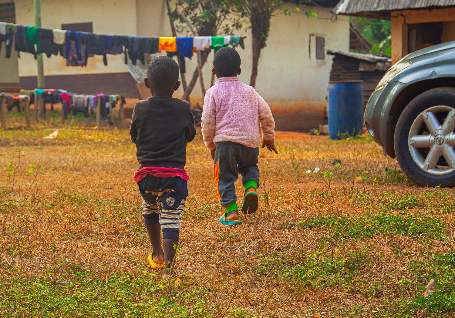 Two young African children running together across a grassy yard near parked cars.
