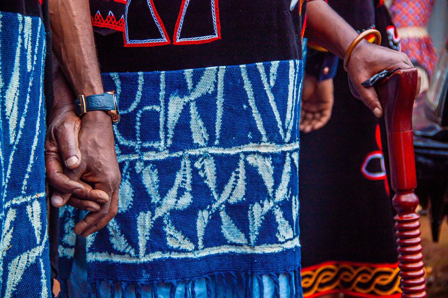 Close-up of hands wearing bracelets, holding a red staff, dressed in traditional blue African batik fabric.