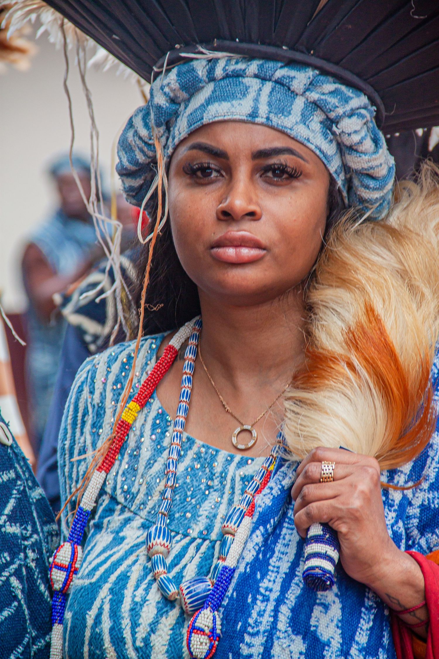 Close-up portrait of African woman in blue traditional dress with beaded necklaces and large feathered hat.