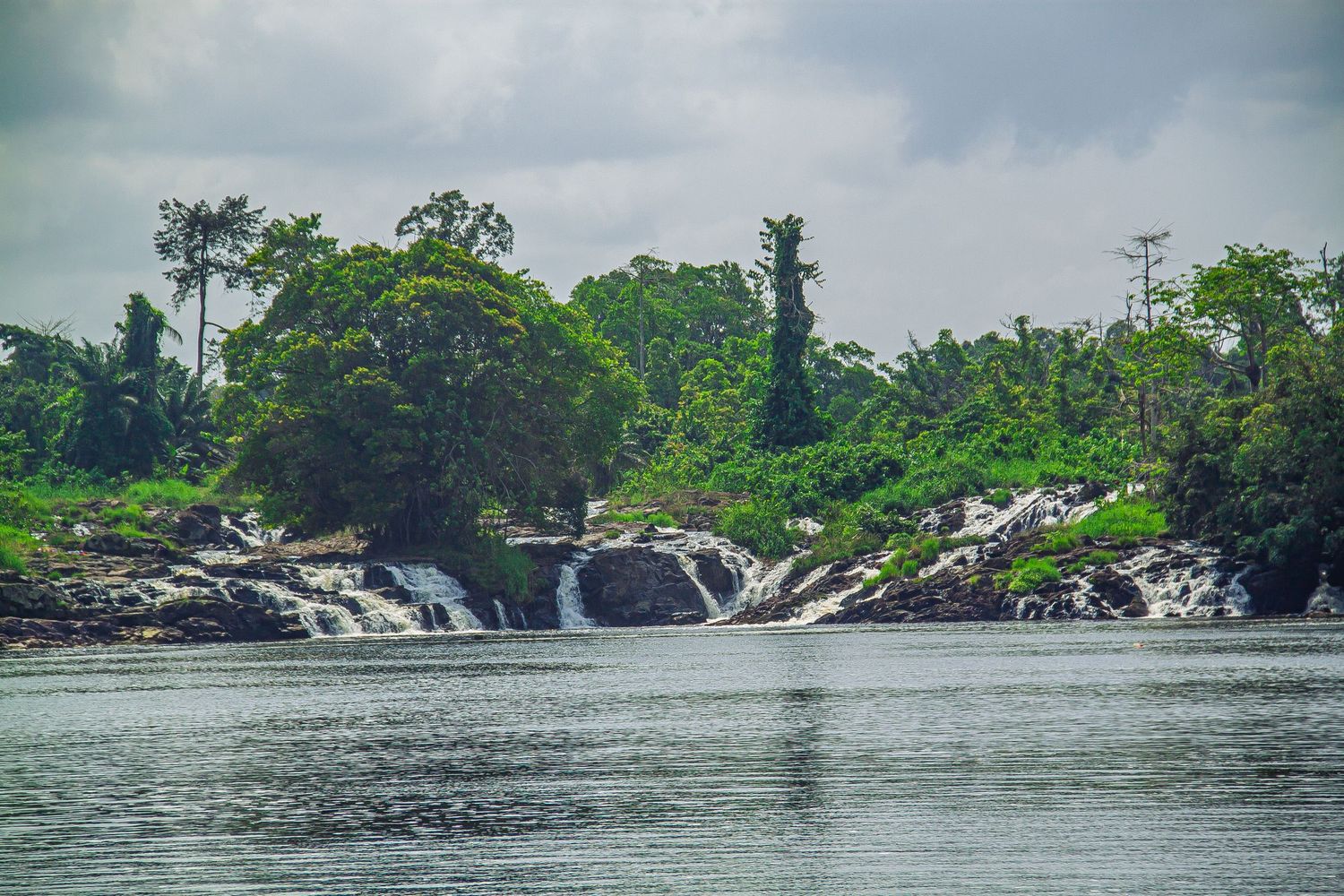A scenic tropical river with cascading waterfalls surrounded by dense green rainforest under a cloudy sky.
