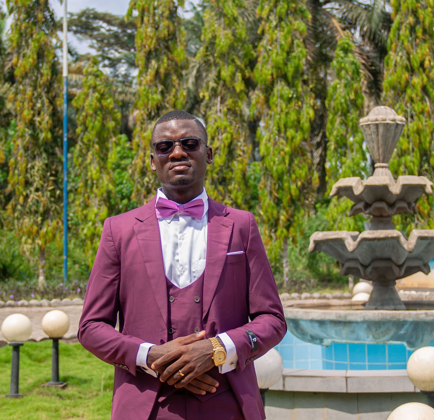 Stylish man in burgundy suit with bow tie posing outdoors near fountain.