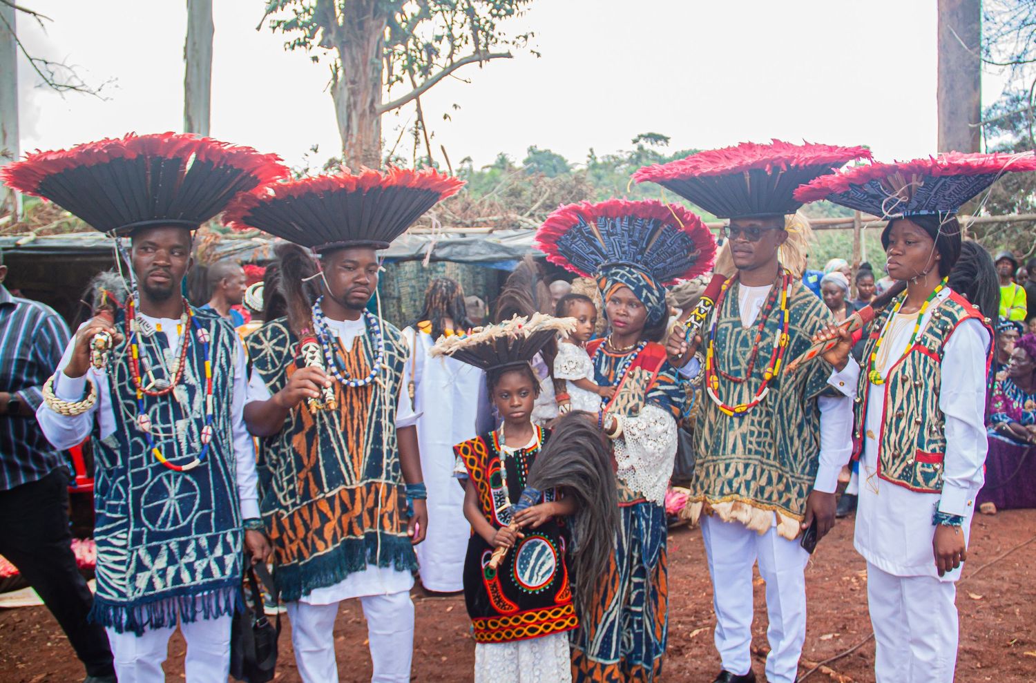 Group of people in colorful traditional African attire with red feathered hats at a cultural festival.